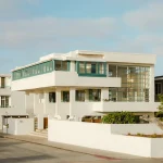 Exterior corner view of the white modernist house with green window frames, surrounding low white walls and neighbouring houses