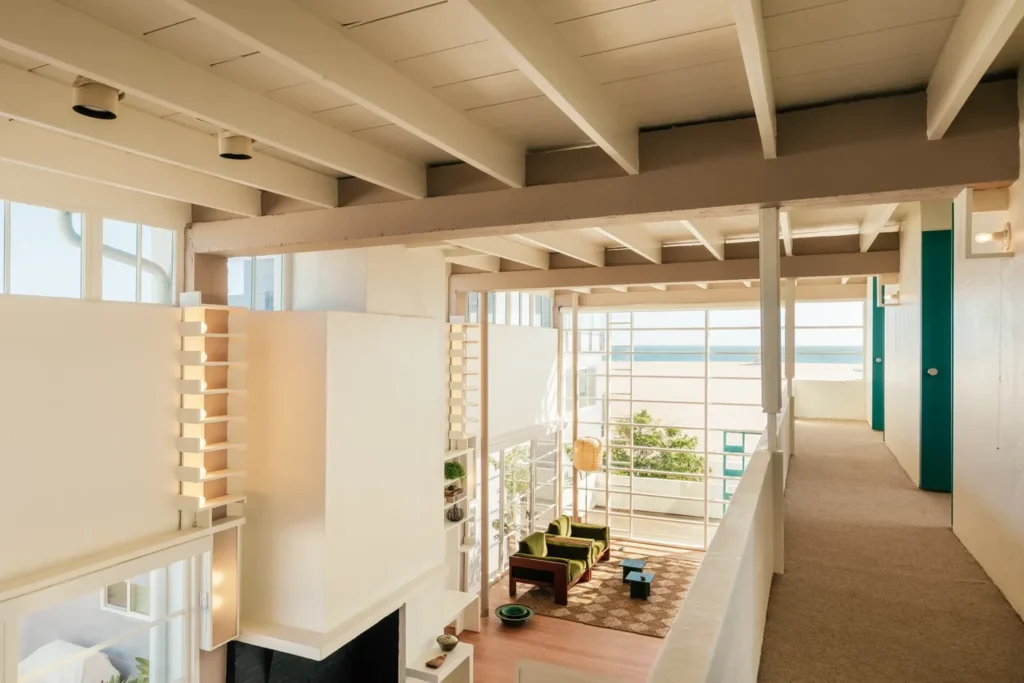 Double-height interior looking toward the ocean through large grid windows, with green seating, built-in shelves and open upper walkway