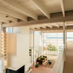 Double-height interior looking toward the ocean through large grid windows, with green seating, built-in shelves and open upper walkway