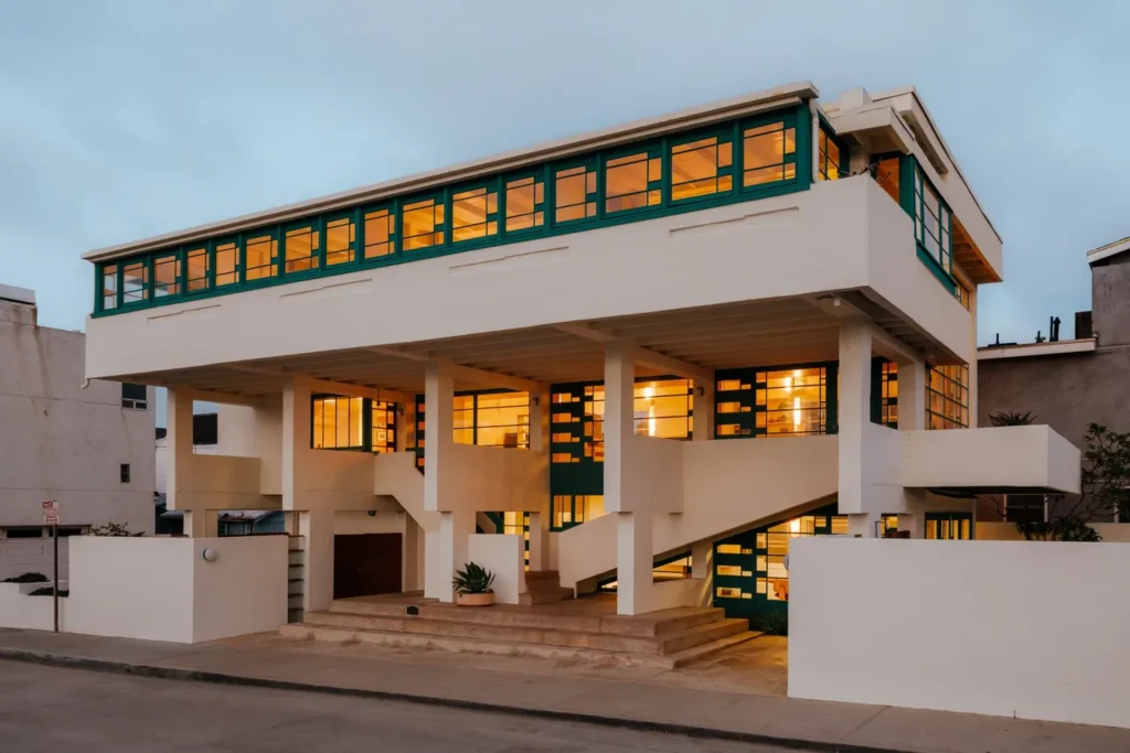 Twilight exterior of the fully illuminated house showing the continuous upper window band, concrete pilotis and open stairs glowing from within
