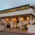 Twilight exterior of the fully illuminated house showing the continuous upper window band, concrete pilotis and open stairs glowing from within