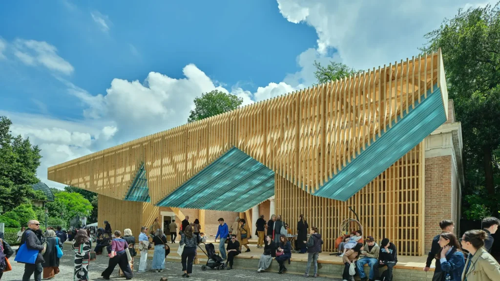 Marlon Blackwell’s Grand PORCH Pavilion to Find a New Home at Crystal Bridges - A timber lattice pavilion with a teal underside canopy installed in front of a classical brick building, surrounded by visitors under a blue sky.