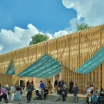 A timber lattice pavilion with a teal underside canopy installed in front of a classical brick building, surrounded by visitors under a blue sky.