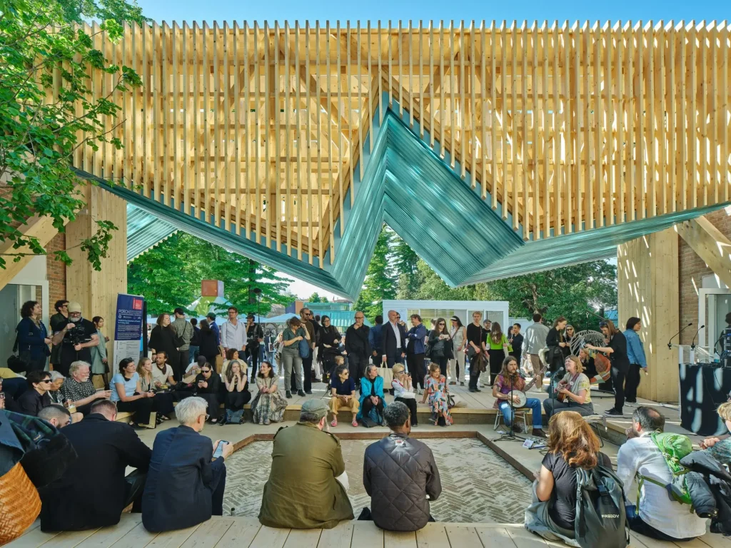 People sitting around a stepped sunken area beneath a folded wooden slatted canopy while live music plays.