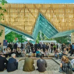 People sitting around a stepped sunken area beneath a folded wooden slatted canopy while live music plays.