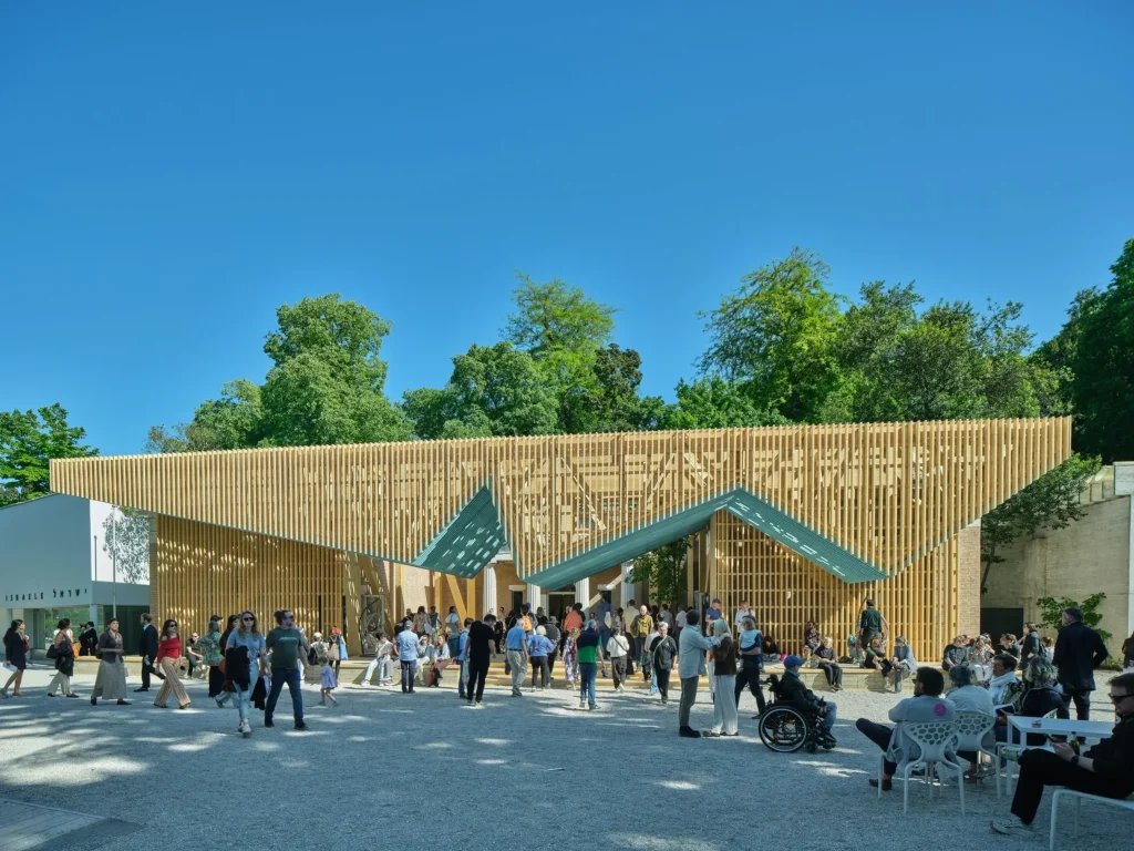 Straight-on front view of a large timber slatted pavilion with dynamic zigzag rooflines spanning the front of a historic building filled with visitors.