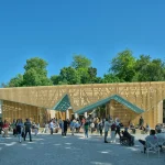Straight-on front view of a large timber slatted pavilion with dynamic zigzag rooflines spanning the front of a historic building filled with visitors.