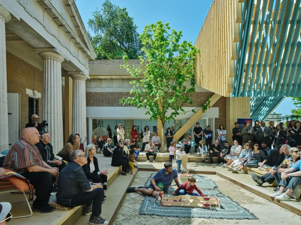 An outdoor courtyard with audience members seated around a central carpeted area where a child plays with model structures under a young tree.