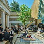 An outdoor courtyard with audience members seated around a central carpeted area where a child plays with model structures under a young tree.