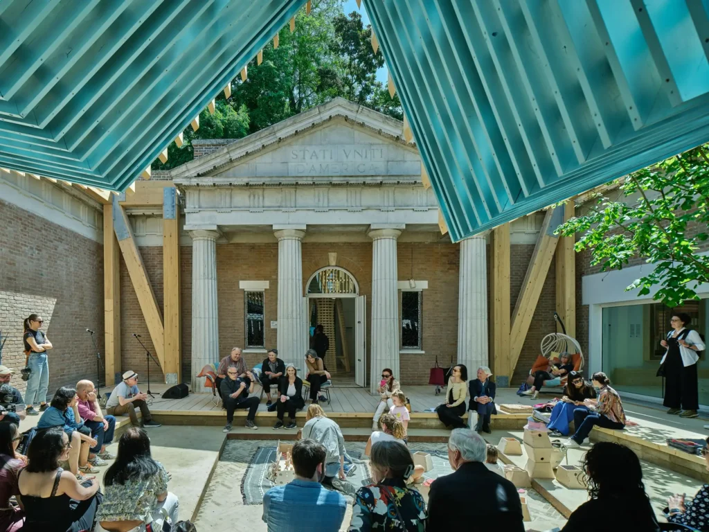Looking up toward a classical building facade with "STATI VNITI D'AMERICA" inscribed, framed by the blue underside of a modern timber roof structure with a seated audience.
