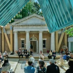 Looking up toward a classical building facade with "STATI VNITI D'AMERICA" inscribed, framed by the blue underside of a modern timber roof structure with a seated audience.