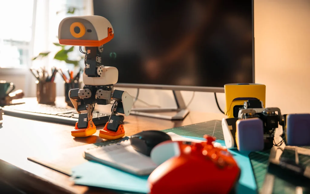 Single white-and-orange Microduck robot standing on a wooden desk beside a computer monitor, keyboard and tools