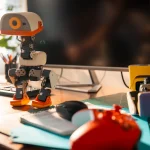 Single white-and-orange Microduck robot standing on a wooden desk beside a computer monitor, keyboard and tools