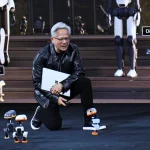 Jensen Huang kneeling on stage holding a laptop and controller, surrounded by several small Microduck robots with larger humanoid robots in the background