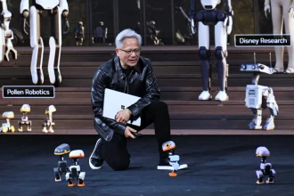 Jensen Huang kneeling on stage holding a laptop and controller, surrounded by several small Microduck robots with larger humanoid robots in the background