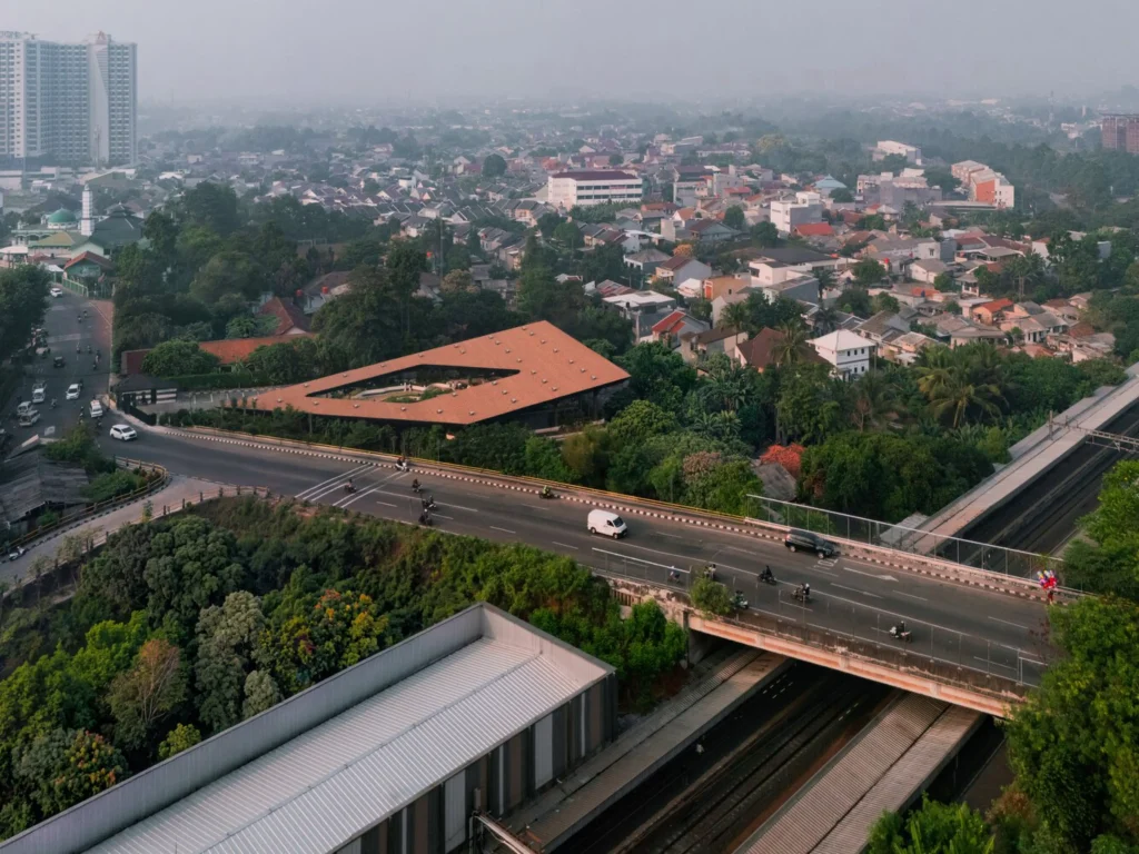 Aerial view of Mitte Oblique Garden in Jakarta showing its large angular terracotta-tiled roof with a central open courtyard, set amid dense urban fabric, a busy elevated highway, railway tracks, and surrounding trees and buildings.