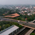 Aerial view of Mitte Oblique Garden in Jakarta showing its large angular terracotta-tiled roof with a central open courtyard, set amid dense urban fabric, a busy elevated highway, railway tracks, and surrounding trees and buildings.