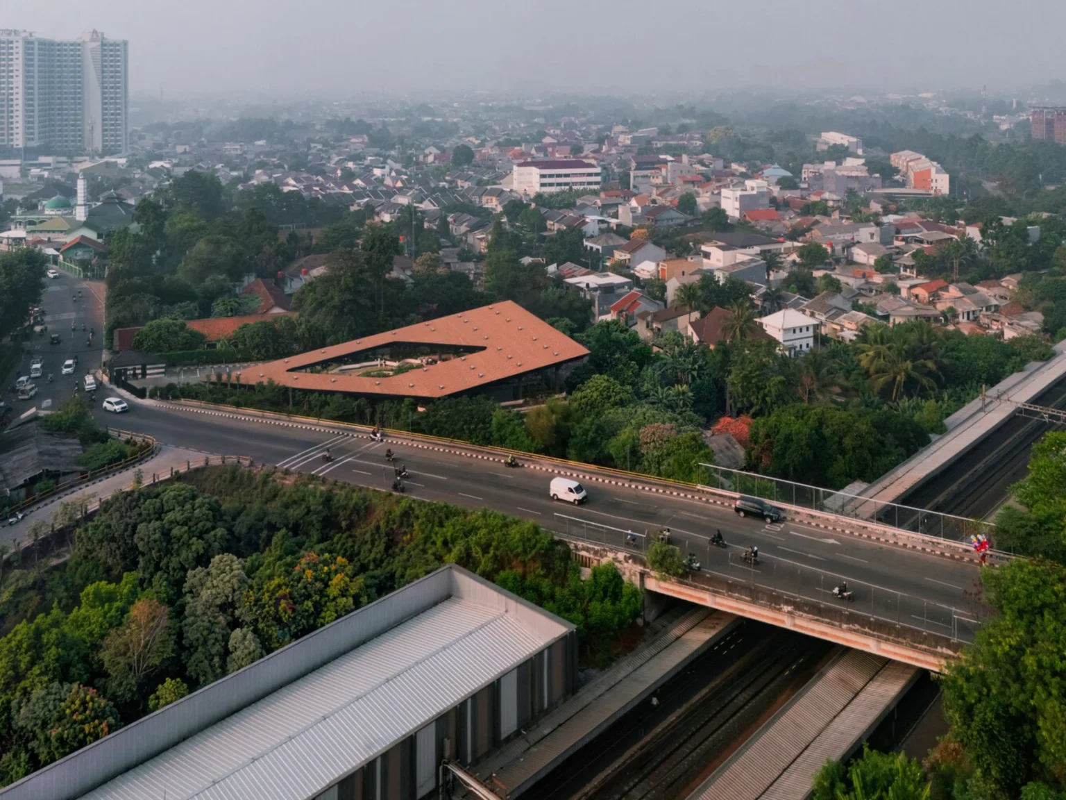 Aerial view of Mitte Oblique Garden in Jakarta showing its large angular terracotta-tiled roof with a central open courtyard, set amid dense urban fabric, a busy elevated highway, railway tracks, and surrounding trees and buildings.