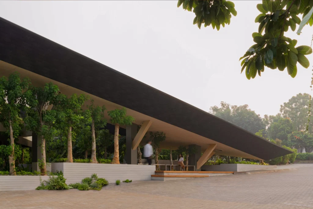 Ground-level view of the deep overhanging dark roof of Mitte Oblique Garden, revealing a shaded outdoor space with trees in raised planters, wooden seating steps, and people walking and resting beneath the canopy.