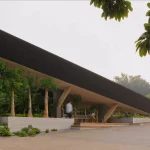 Ground-level view of the deep overhanging dark roof of Mitte Oblique Garden, revealing a shaded outdoor space with trees in raised planters, wooden seating steps, and people walking and resting beneath the canopy.