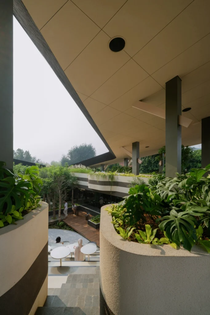 View from under the canopy looking toward the open courtyard of Mitte Oblique Garden, with large concrete planters filled with tropical plants, a wooden deck, seating areas, and people occupying the multi-level space.