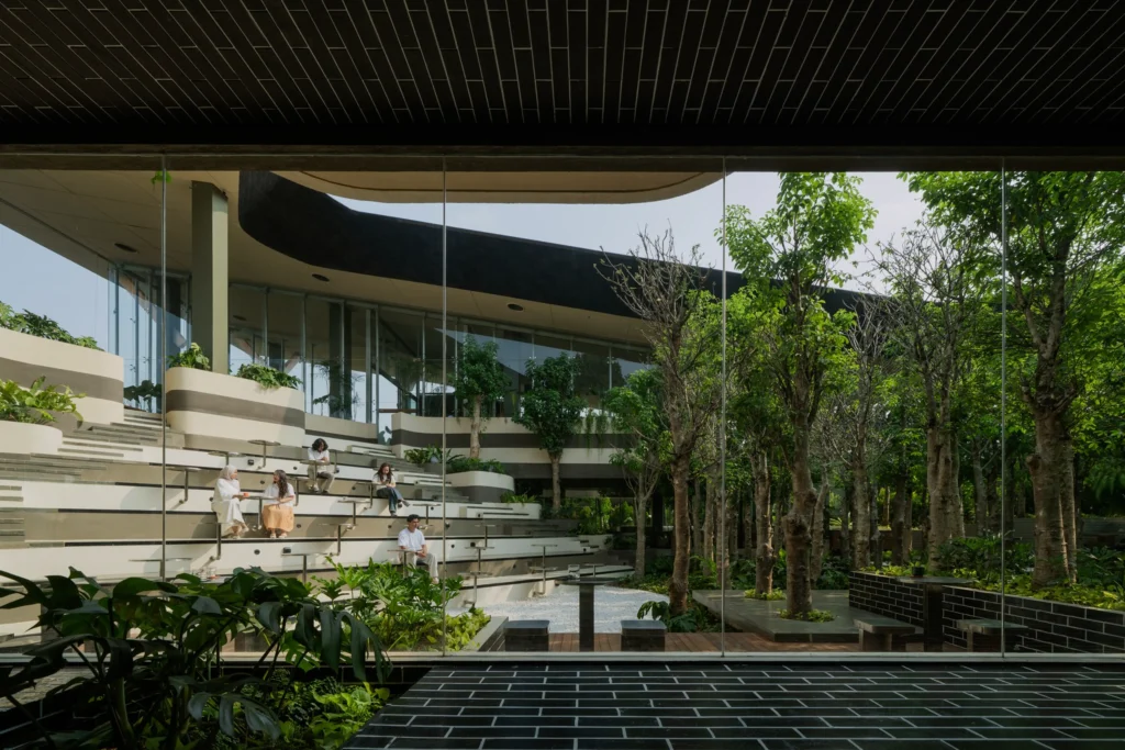 Wide view through glass walls of the multi-level amphitheater-style seating and dense tree plantings inside Mitte Oblique Garden, with people sitting on the stepped concrete and wood platforms.