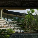 Wide view through glass walls of the multi-level amphitheater-style seating and dense tree plantings inside Mitte Oblique Garden, with people sitting on the stepped concrete and wood platforms.