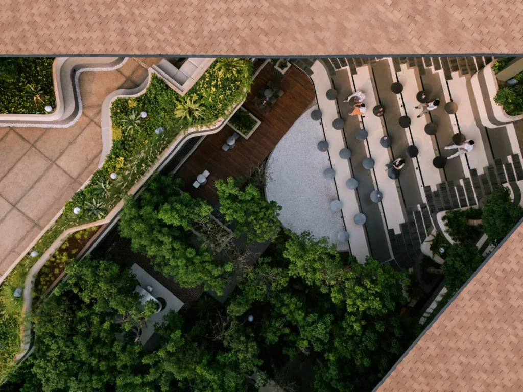 Bird’s-eye view looking straight down into the central courtyard of Mitte Oblique Garden, showing curved planters, wooden decks, circular tables, gravel areas, and dense tree canopy framed by the terracotta roof edges.