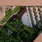 Bird’s-eye view looking straight down into the central courtyard of Mitte Oblique Garden, showing curved planters, wooden decks, circular tables, gravel areas, and dense tree canopy framed by the terracotta roof edges.