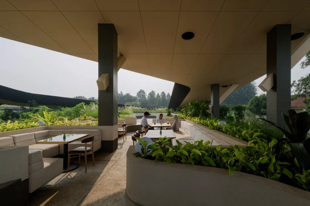 Covered outdoor dining and lounge area at Mitte Oblique Garden with curved concrete seating, tables, dense surrounding greenery, and people seated under the large canopy.