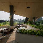 Covered outdoor dining and lounge area at Mitte Oblique Garden with curved concrete seating, tables, dense surrounding greenery, and people seated under the large canopy.