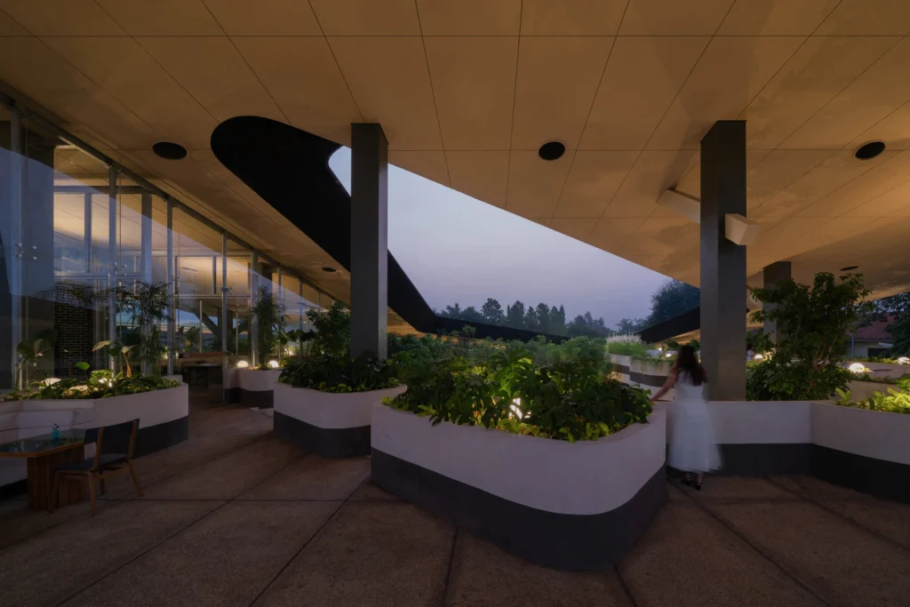 Dusk view of the covered outdoor spaces at Mitte Oblique Garden, with illuminated planters, curved seating, glass walls, and a person standing near the edge of the open roof void.