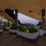 Dusk view of the covered outdoor spaces at Mitte Oblique Garden, with illuminated planters, curved seating, glass walls, and a person standing near the edge of the open roof void.