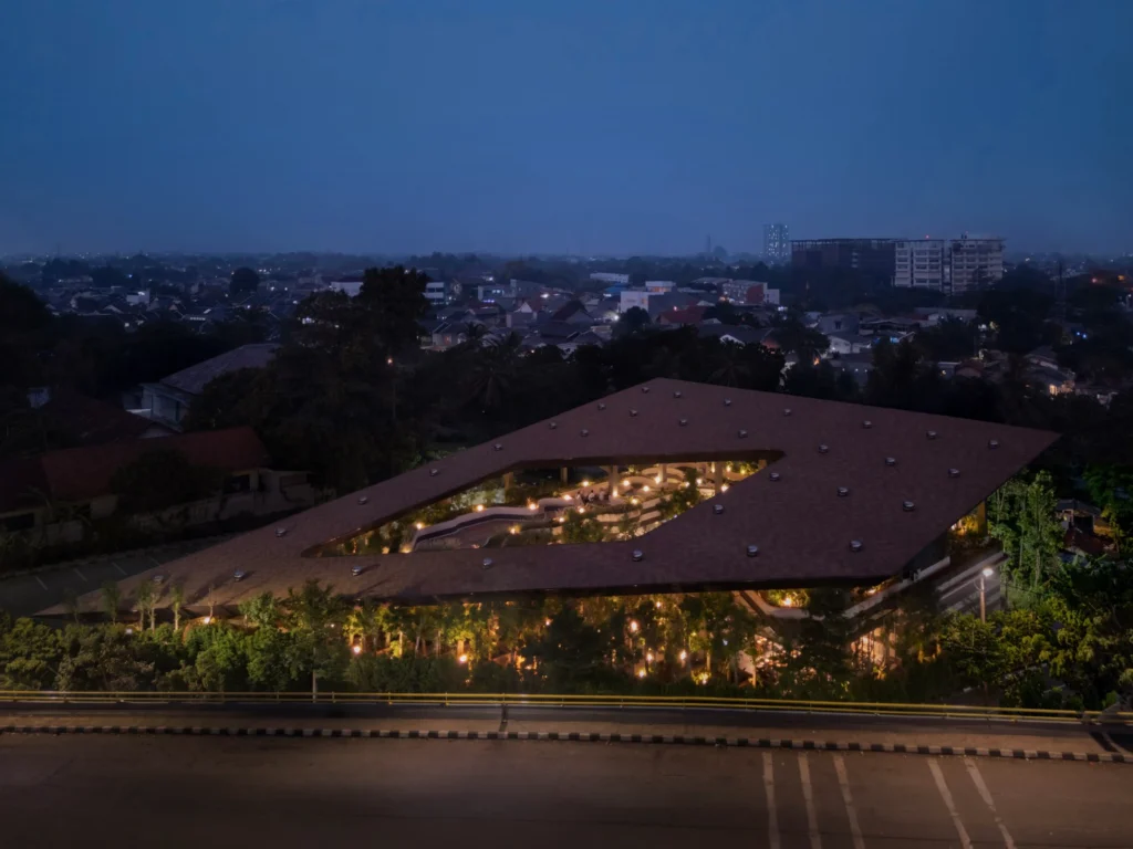 Night aerial view of Mitte Oblique Garden showing its illuminated angular roof and glowing interior courtyard against the dark urban landscape of Jakarta.