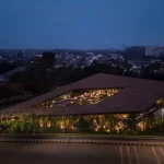 Night aerial view of Mitte Oblique Garden showing its illuminated angular roof and glowing interior courtyard against the dark urban landscape of Jakarta.