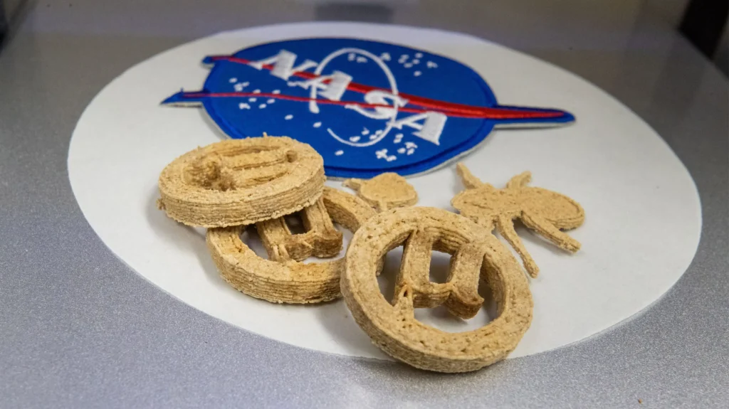 Three golden-brown 3D-printed µBites cookies arranged on a white surface with a soft blue NASA logo in the background