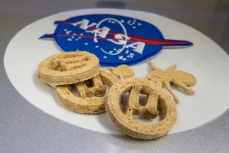 Three golden-brown 3D-printed µBites cookies arranged on a white surface with a soft blue NASA logo in the background