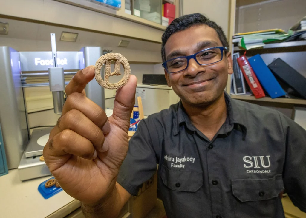Researcher in an SIU shirt smiling and holding up a translucent 3D-printed cookie in a laboratory