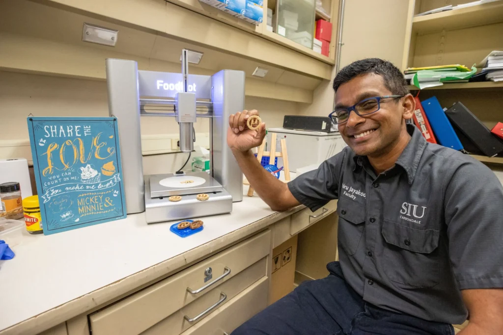 Researcher in an SIU shirt smiling and holding up a translucent 3D-printed cookie in a laboratory