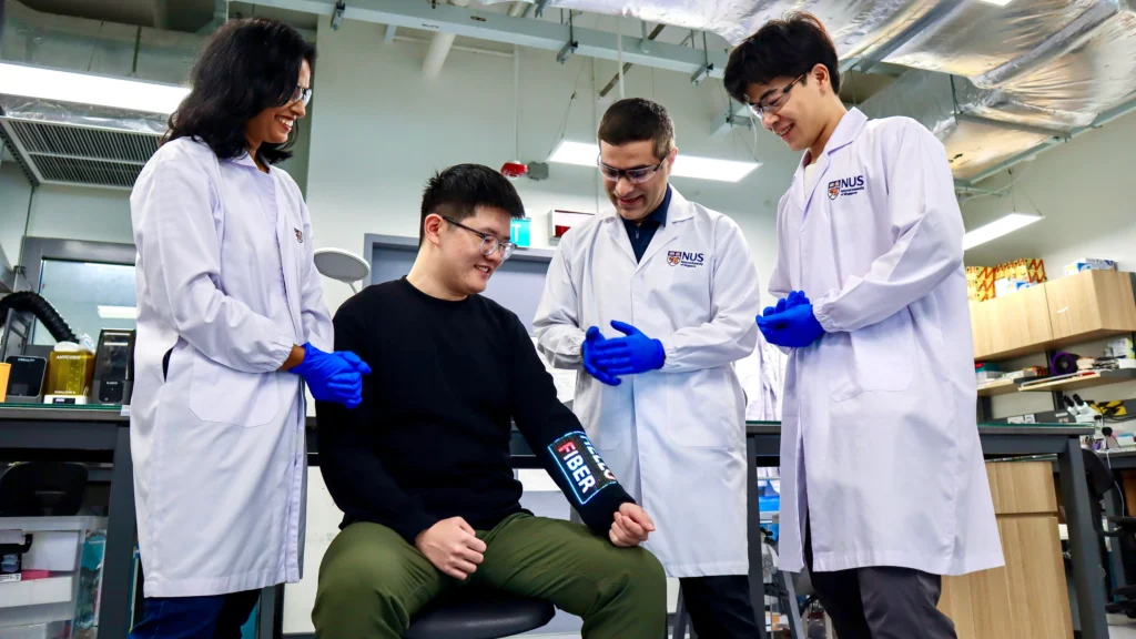Four researchers in white lab coats and blue gloves gather around a smiling team member wearing a black sleeve with an electronic fiber display on the forearm