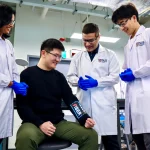 Four researchers in white lab coats and blue gloves gather around a smiling team member wearing a black sleeve with an illuminated fibre display on the forearm