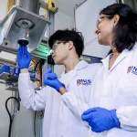 Two researchers in white NUS lab coats and blue gloves examine a thin electronic fibre being drawn from laboratory equipment