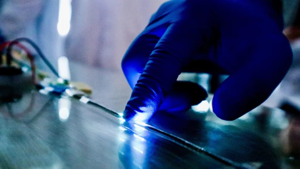 Close-up of a gloved hand pressing a thin electronic fibre that lights up brightly under blue laboratory lighting