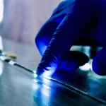 Close-up of a gloved hand pressing a thin electronic fibre that lights up brightly under blue laboratory lighting