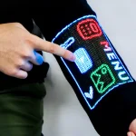 Close-up of a hand tapping a glowing menu interface displayed on the black sleeve, showing colourful icons including a calculator and temperature symbol