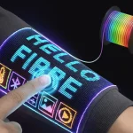 Close-up of a finger pressing a glowing touchscreen woven into a dark sleeve, displaying the word “FIBER” in blue pixels along with colourful icons for temperature, photos and media, while a spool of rainbow electronic fibre floats nearby