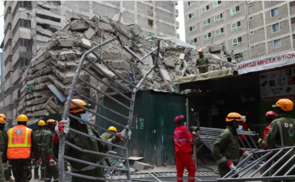 Emergency workers in orange and yellow helmets clearing metal debris near a green shop front as the collapsed concrete mound rises behind them