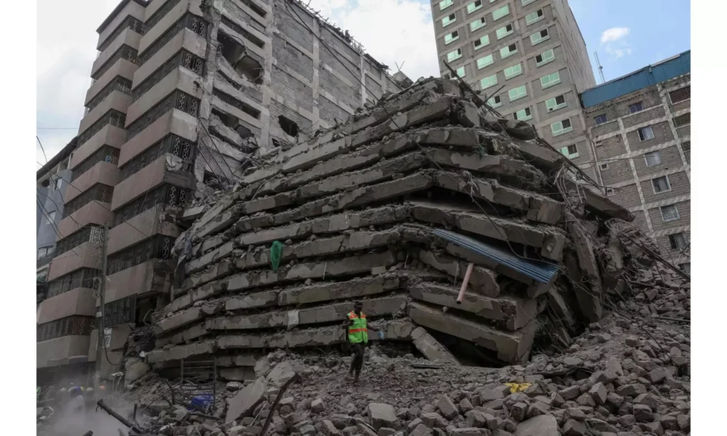 Massive pile of collapsed concrete floor slabs stacked like pancakes beside damaged multi-storey buildings, with a worker in a yellow safety vest standing in the foreground