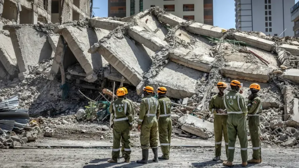 Group of rescue workers in green uniforms and orange helmets standing in front of twisted concrete slabs from the collapsed building
