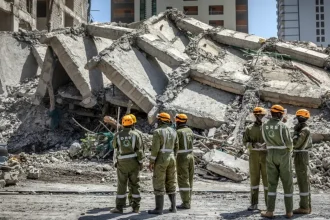 Group of rescue workers in green uniforms and orange helmets standing in front of twisted concrete slabs from the collapsed building